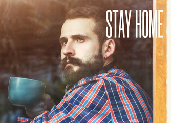 bearded man sitting with cup of morning coffee or tea near window with reflection