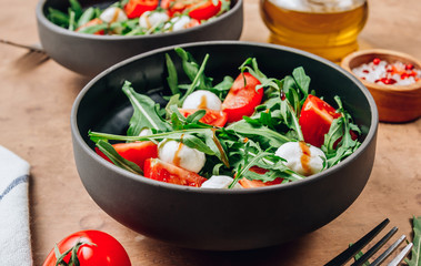 Healthy arugula salad with mozzarella cheese and cherry tomatoes in black bowls on beige rustic background.