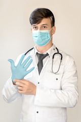 Theme of health and healthcare. A young doctor with dark hair stands in a white coat and a mask from the virus, with a stethostop, wears blue gloves. vertical frame