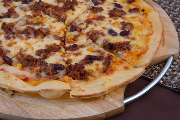 Pizza with lots of cheese and minced meat, sweet pickled corn, raisins and a little tomato on a wooden plate on a brown background close-up