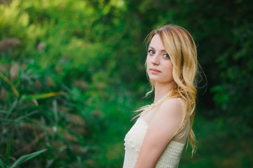 Naklejka premium young, blonde girl, of Slavic origin, in a white dress of the bride against the background of summer green nature. A girl in a white dress among green trees in the forest