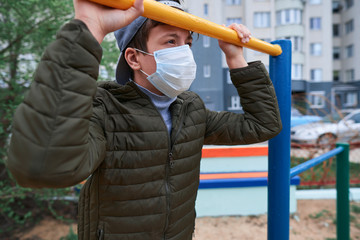 teen boy stands near horizontal bar of outdoor workout equipment, on playground near high-rise buildings with apartments, a medical mask on his face protects against viruses and dust