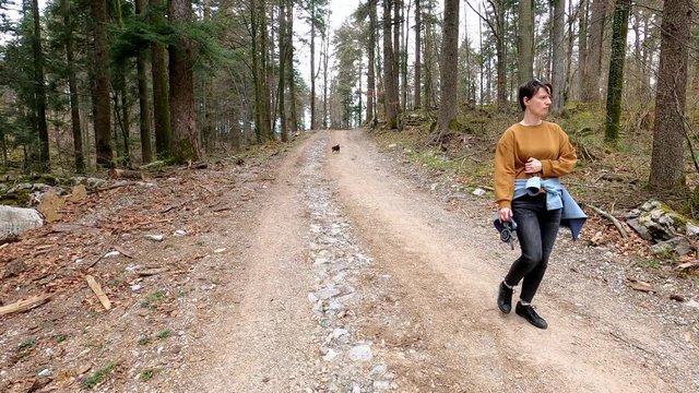 Woman in casual clothes walking on the forest macadam trail, small black dog following her, footage with action camera
