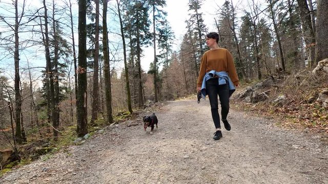 Woman in casual clothes walking on the forest macadam trail, small black dog following her, footage with action camera
