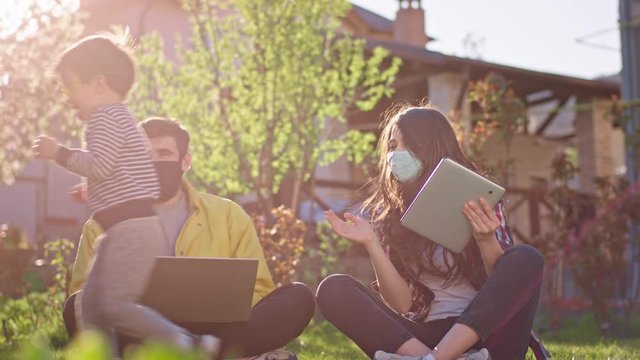 Concentrated working parents online from home while sitting down on the grass they using laptop and tablet to work their small son playing at them quarantine time stay home be safe wearing protective