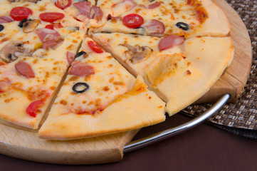 Big cheese pizza with tomatoes, mushrooms and smoked sausage on a wooden round board close-up on a brown background