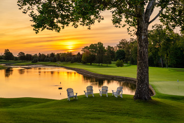 Peaceful and tranquil Orange and yellow sunrise over a  pristine lake at Golfcourse with Adirondack chairs in the foreground.