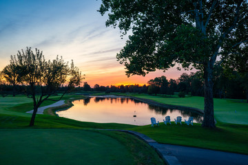 Peaceful and tranquil sunrise over a small body of pristine smooth water at Golfcourse with Adirondack chairs in the foreground.