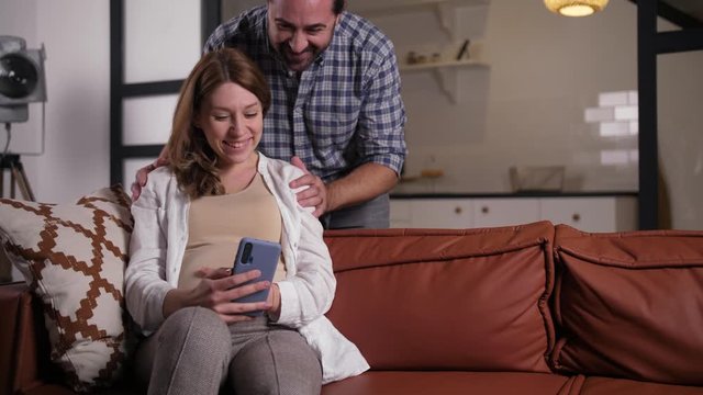 Closeup of cheerful couple expecting baby communicating online with relatives via internet from home. Smiling pregnant woman sitting on sofa with smartphone, loving husband hugging her, stroking tummy