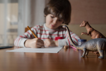 Preschooler drawing on the table in his home with dinosaurs toys