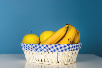 Ripe fruit bananas and apples in white basket on the table in front of the blue wall background in day organic food vegan or vegetarian