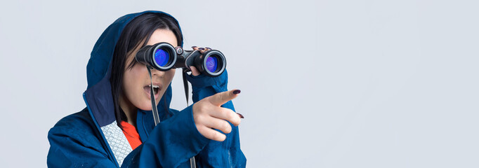 A tourist girl in a blue raincoat holds binoculars in her hands and looks into the distance, spies.