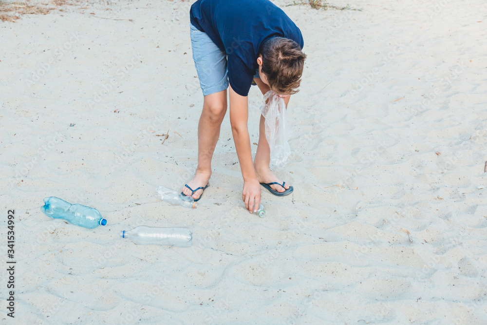 Wall mural boy in white t shirt in gloves collects garbage and plastic bottles into package on the beach. young