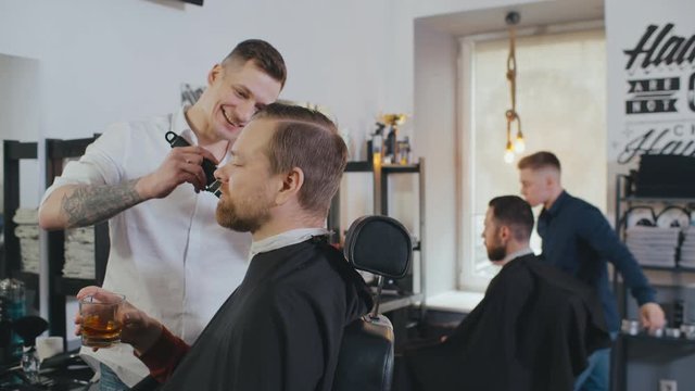Handsome client drinking whiskey while barber trimming his hair