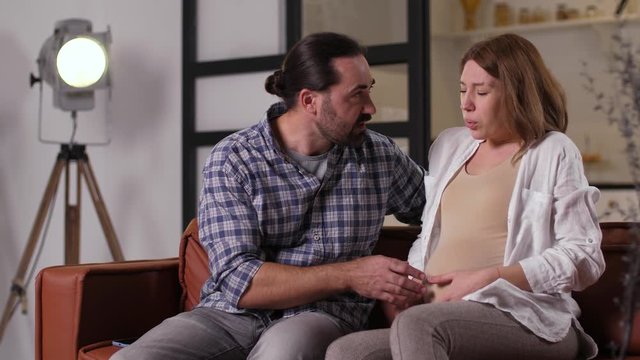 Closeup of scared pregnant couple during conctractions of childbirth. Frightened woman sitting on sofa during start of labor pains, confused husband taking cellphone and calling 911 ambulance