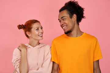 Glad young ginger female and dark skinned male in casual clothes, look with positive expressions, enjoy spare time together, people over pink wall, have friendly relationships. Two people indoor