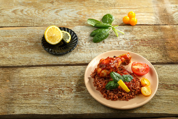 Chicken legs with red rice in tomato-garlic sauce on a plate on a wooden table near lemon and spinach.