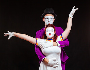 Portrait of two mime artists, isolated on black background. Young woman stands with risen hands, man hugs her from behind, looking at camera