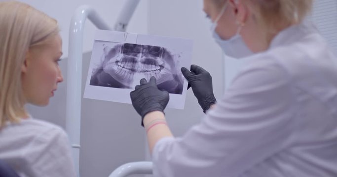Dentist doctor looks with the patient an x-ray picture of the oral cavity. Dentist shows on an x-ray which teeth should be treated. Modern dental office with professional equipment.