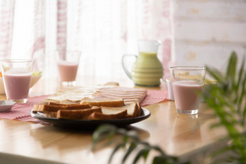 Healthy breakfast served on wooden table in space with window and natural light with yogurt and green smoothie in glasses and glass jar, chopped bread on black plate, ham and cheese on wooden table