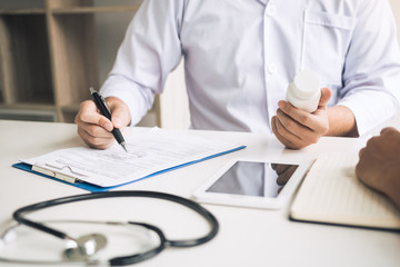 Confident doctor man holding a pill bottle and writing while talking with senior patient and reviewing his medication at office room.