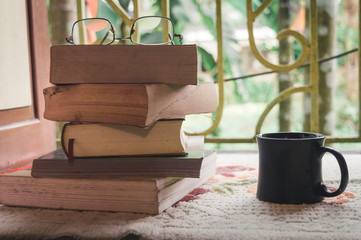 Obraz premium A black cup of coffee and a heap of books and reading glass on window sunlight in morning during social distancing. Stay Entertained at Home Holiday leisure Education and Healthy lifestyle background.