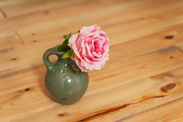 Pink rose in a green jug on the background of a wooden table. One blooming rosebud in a vase on the...