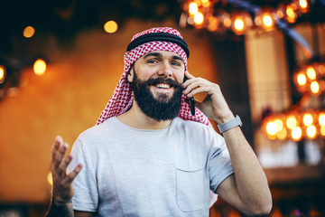 Attractive smiling tattooed bearded trendy muslim guy sitting in cafe in the morning and having a peasant conversation with his friend on mobile phone.