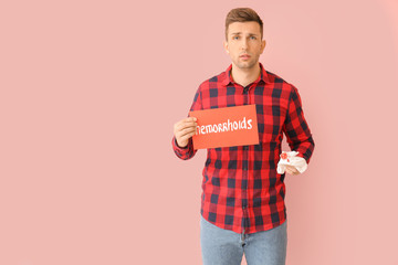 Young man holding paper with text HEMORRHOIDS and toilet paper in blood on color background