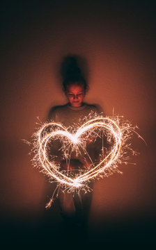 Girl Making Heart Shape With Sparkler In Darkroom Against Wall
