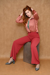 The model is wearing a pink shirt and red pants. Vertical portrait full length sitting on a stand Caucasian red hair pretty woman on a beige background.