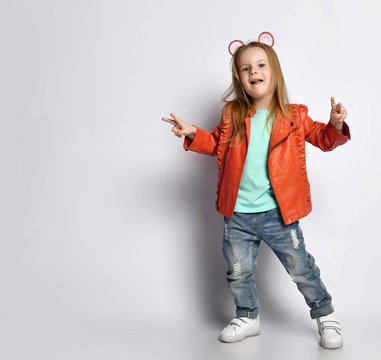Playful blond baby girl kid in leather jacket, white t-shirt and blue jeans gestures V-signs victory 