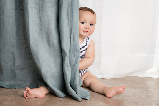 Cute baby sitting on floor peeking behind curtains at home