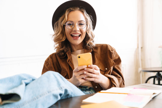Image of smiling young woman using cellphone with legs on table
