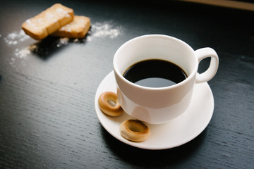 Simple set up for morniing coffee break which includes coffee cup and saucer and few biscuits and bagels plus a bunch of cinnamon sticks. and sugar powder for decoration. black background
