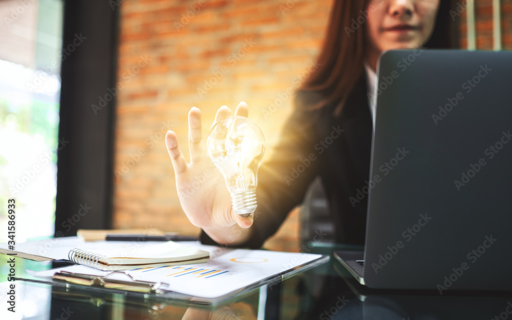 Wall mural closeup image of a businesswoman holding a glowing light bulb while working on laptop computer in of