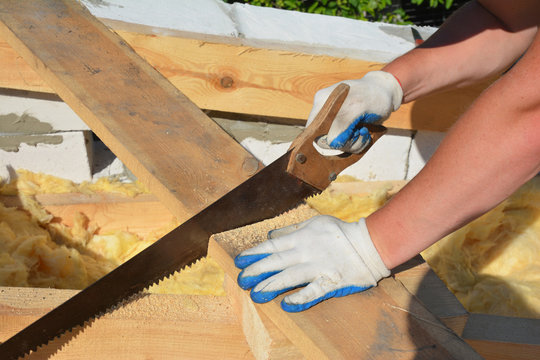 A building contractor in protective gloves is sawing a wooden plank, board with a hand saw to frame the rooftop as one of the stage in roofing construction.