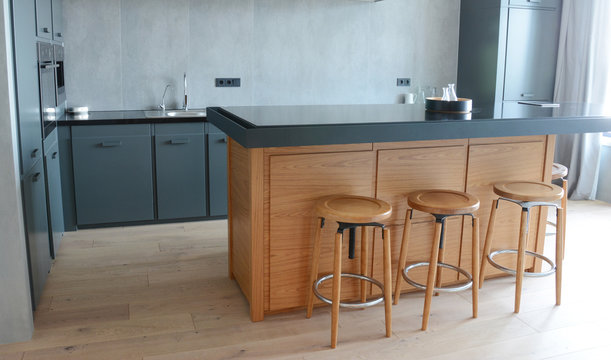 A modern kitchen with dark gray kitchen countertop with a metal sink with steel faucet, wall mounted electrical plugs, cabinets and wooden kitchen island with brown bar stools on laminate wood floor.