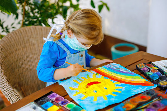 little toddler girl in medical mask painting rainbow with water colors during pandemic coronavirus quarantine disease. Children painting rainbows around the world with the words Let's all be well.