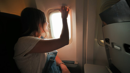 The girl closes and opens the curtain of the illuminator on the plane and enjoys the flight.