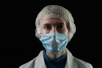 Young female doctor in medical mask, medical gown and cap on black background with copy space