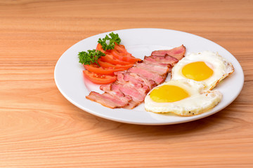 Breakfast with bacon, fried egg and tomatoes on served on white plate over light rustic wooden table.
