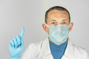 Male doctor in a medical gown and a medical mask stands on a light background with a raised finger up with copy space.