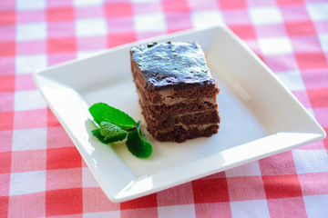 Chocolate brownie piece on white plate decorated with mint leaves served on a white plate over red plaid tablecloth.