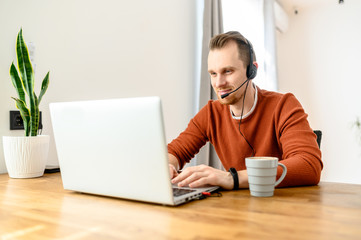 The guy uses hands-free headsets to work from home. He sits at a table with a laptop and speaks online. Remote work concept