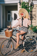 Beautiful blond woman in large wide-brimmed hat riding a bicycle in a town with her cute brown dog breed french bulldog which sits in a wooden basket of a retro bicycle, traveling with a dog, concept