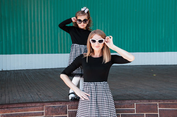 Young beautiful mom with her daughter posing against wall, minimalist street fashion style, family...
