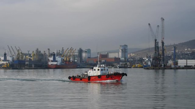 The red tugboat is sailing in the water area of the industrial city against the background of the port