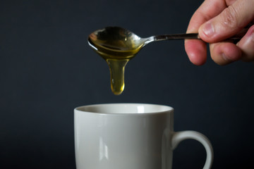 Close up of hand pouring honey from spoon to white cup, mug in front of black background