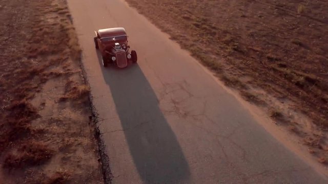 Drone Aerial View Classic Car Hot Rod Driving Along Old Abandoned Road At Sunset Casting Shadow